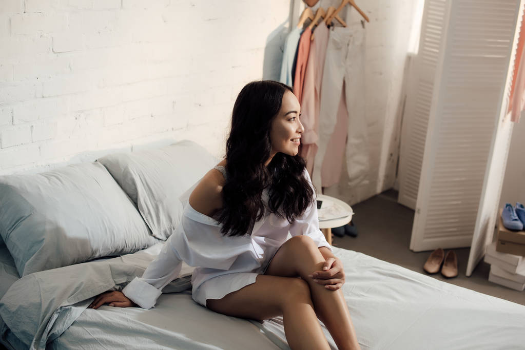 seducente sorridente asiatico ragazza seduta su letto e guardando lontano a casa
 - Foto, immagini