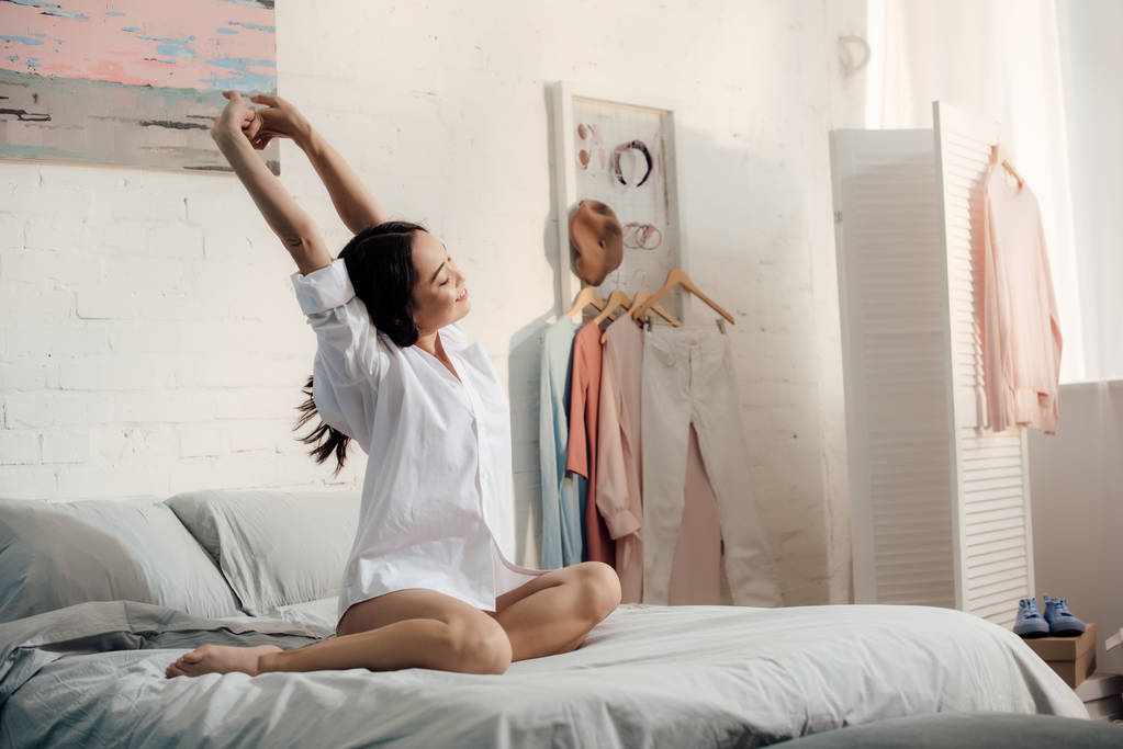 привлекательная азиатская женщина в белой рубашке сидит на кровати и протягивает руки утром
 - Фото, изображение