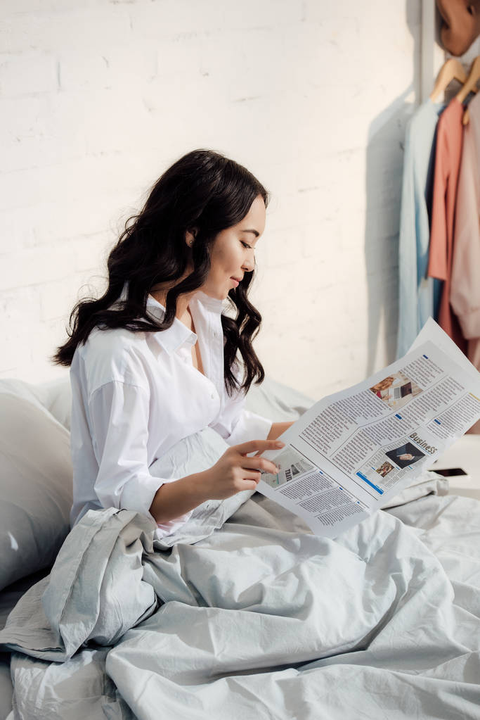 όμορφη νεαρή γυναίκα της Ασίας διαβάζοντας εφημερίδα ενώ κάθεται στο κρεβάτι το πρωί - Φωτογραφία, εικόνα