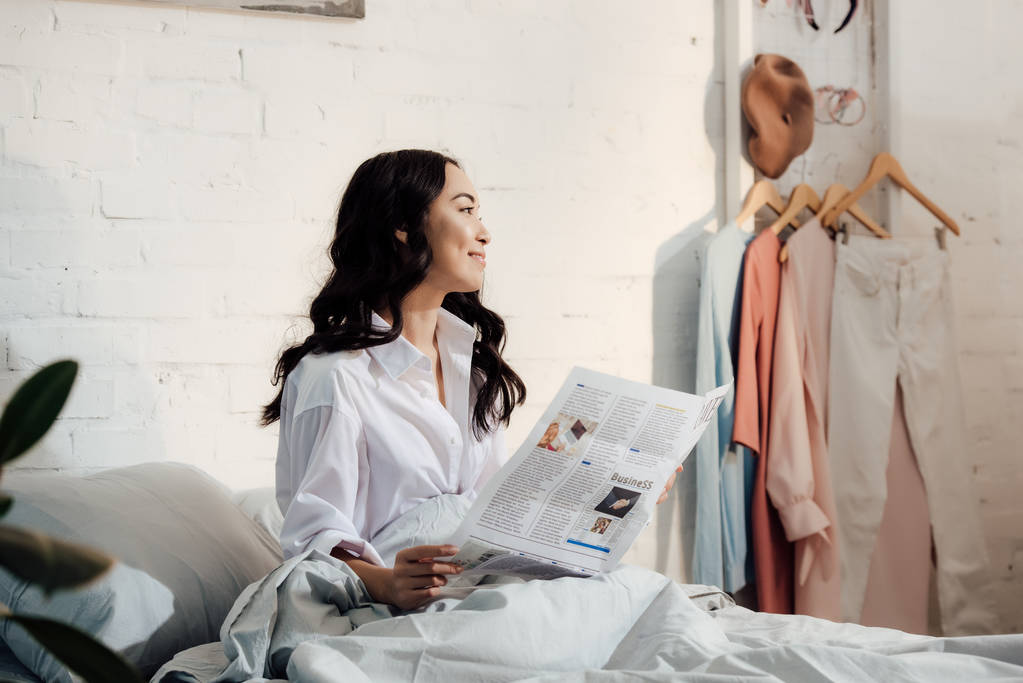 mooi lachende meisje in wit overhemd krant te houden en weg te kijken in de slaapkamer - Foto, afbeelding
