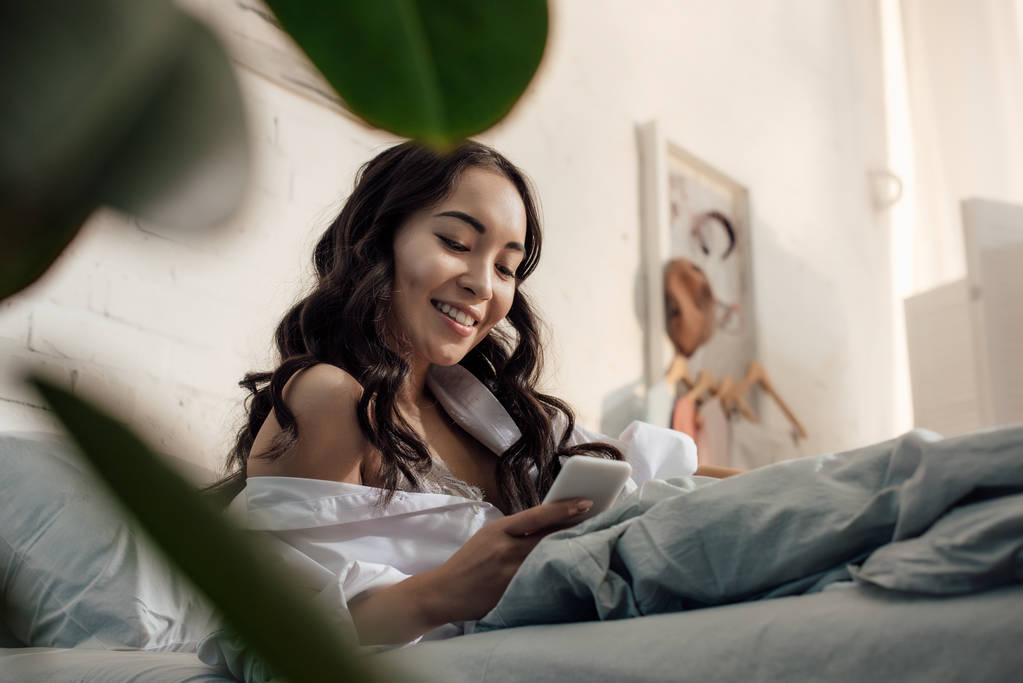 низкий угол зрения красивой улыбающейся азиатской девушки с помощью смартфона в постели
 - Фото, изображение