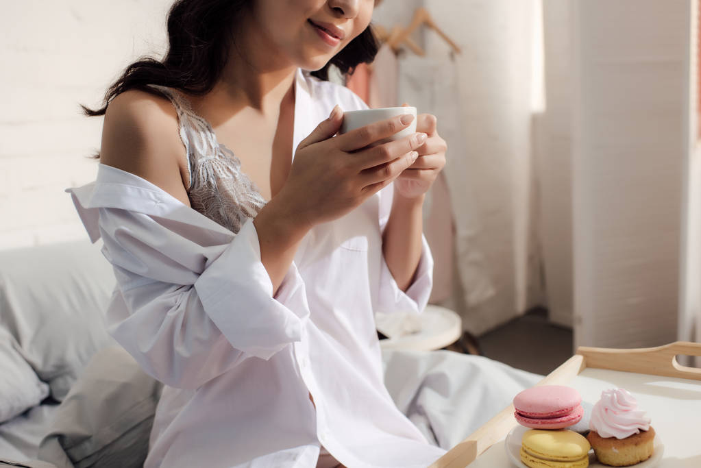 обрезанный снимок улыбающейся азиатской девушки в лифчике и белой рубашке, держащей чашку кофе и сидящей на кровати
 - Фото, изображение