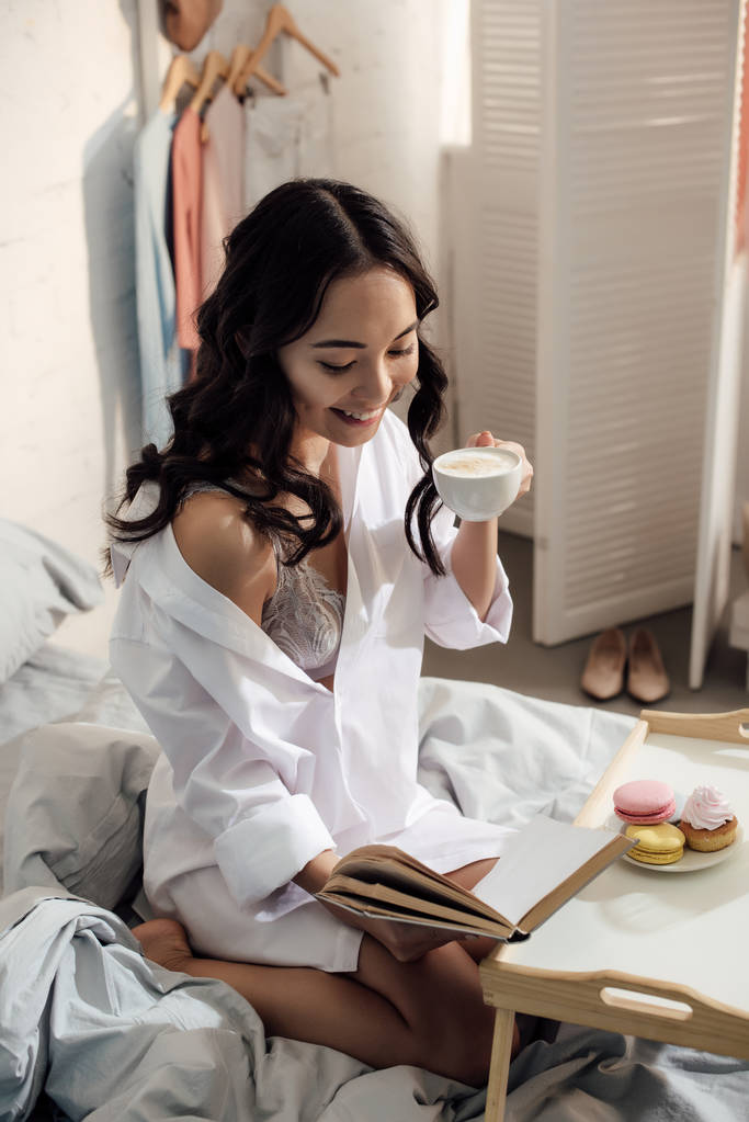 beautiful smiling asian girl reading book and drinking coffee on bed - Photo, Image