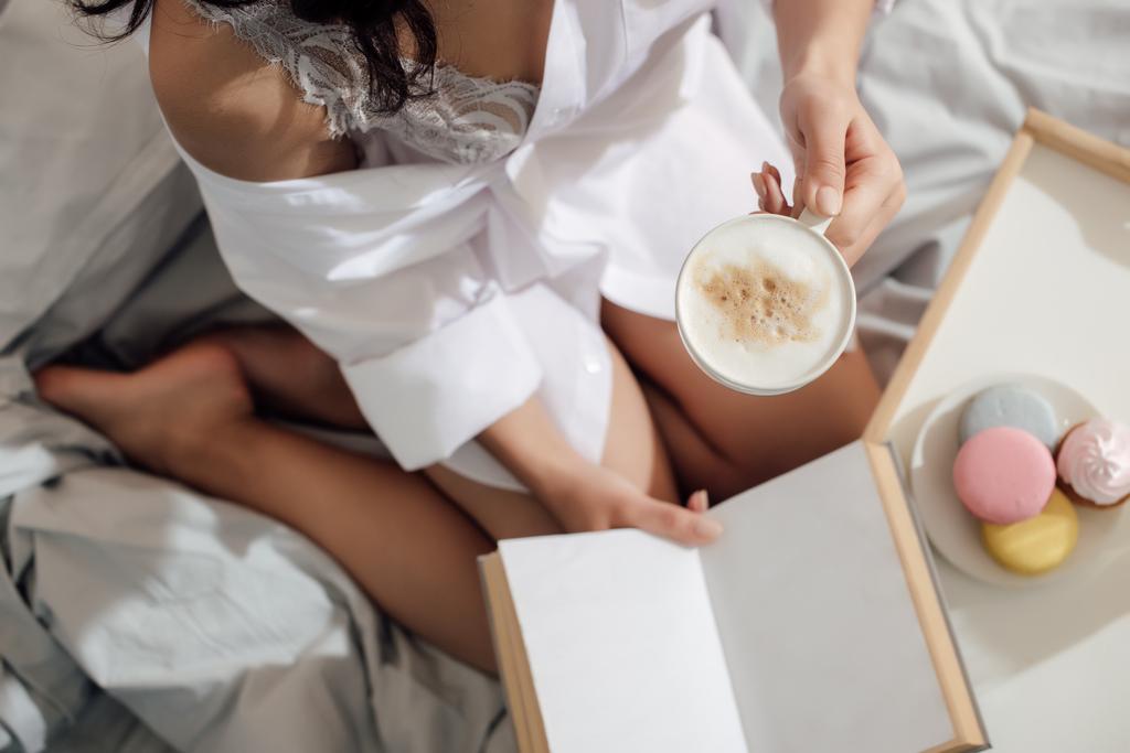 Schnappschuss eines verführerischen Mädchens, das eine Tasse Cappuccino in der Hand hält und Buch auf dem Bett liest - Foto, Bild