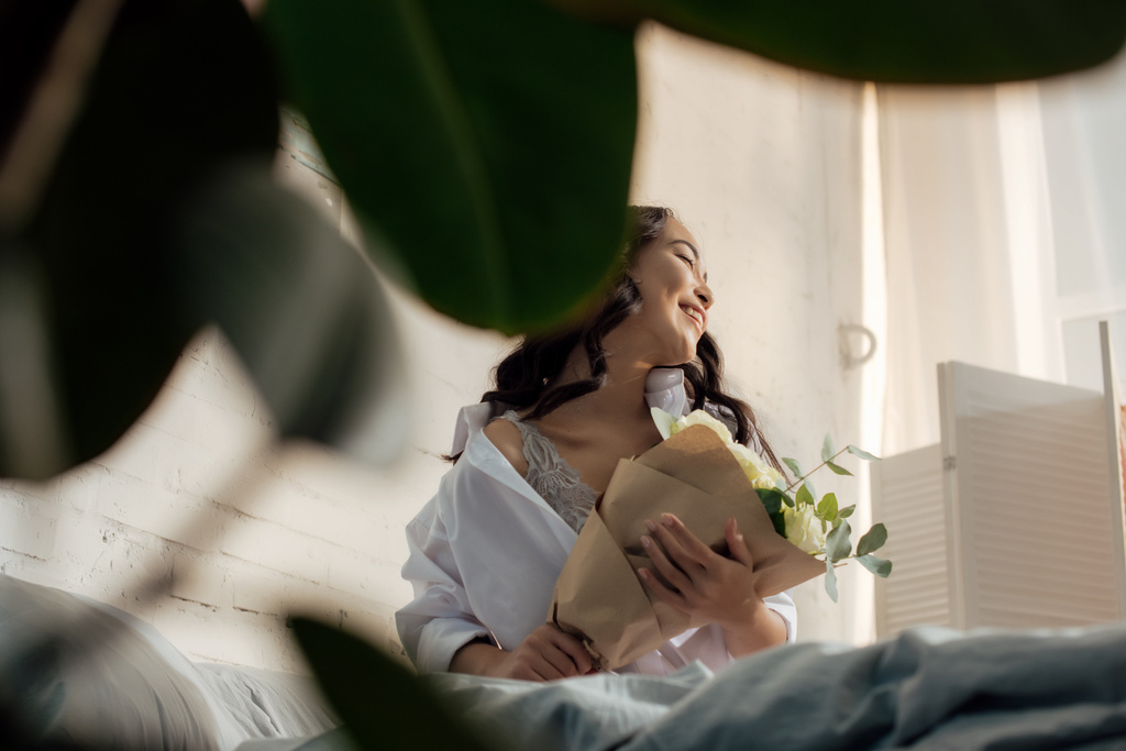 バラの花束が付いているベッドの上に座って白いシャツで美しい幸せな若いアジア女性の選択と集中  - 写真・画像