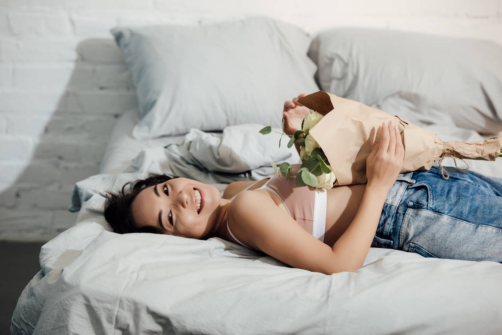 Gelukkig jonge vrouw houdt van mooie bloemboeket en glimlachend in de camera terwijl liggend op bed - Foto, afbeelding