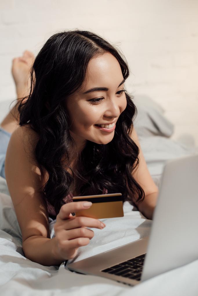 güzel gülümseyen kız yatakta yatarken ve laptop ve kredi kartı ile alışveriş online - Fotoğraf, Görsel