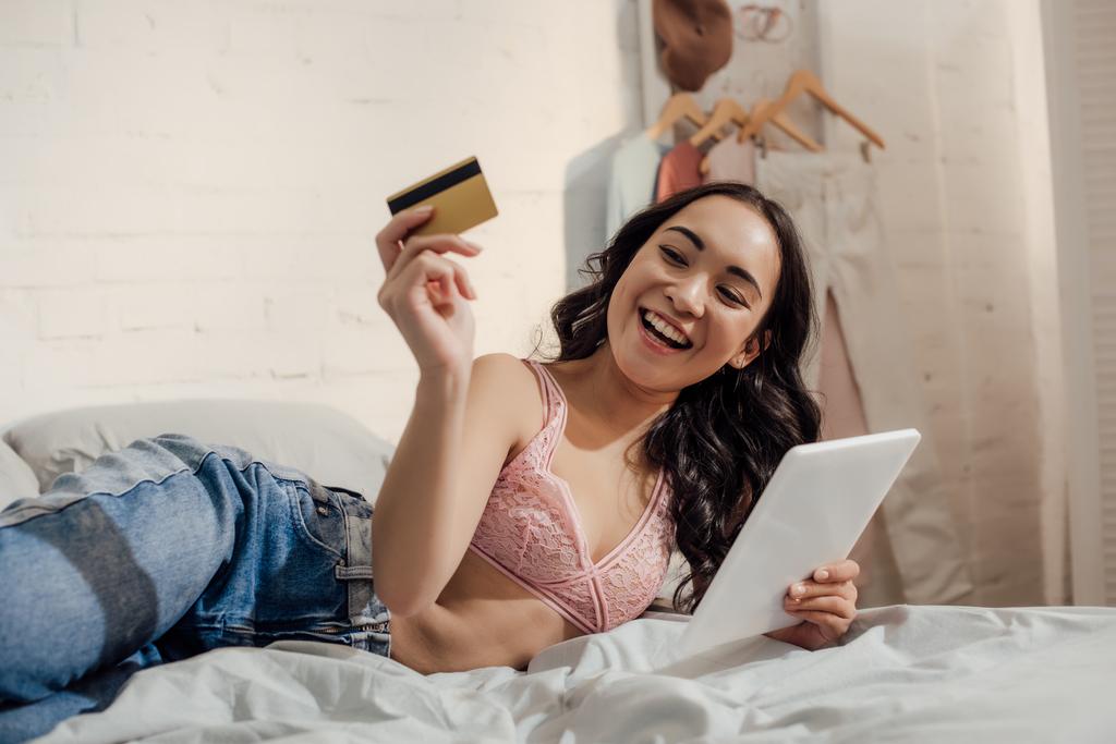 alegre joven de compras en línea con tableta digital y tarjeta de crédito en el dormitorio
  - Foto, imagen