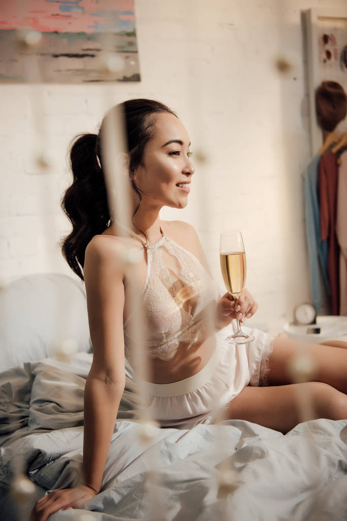 hermosa sonriente chica asiática en ropa interior sentado en la cama con copa de champán y mirando hacia otro lado
 - Foto, Imagen