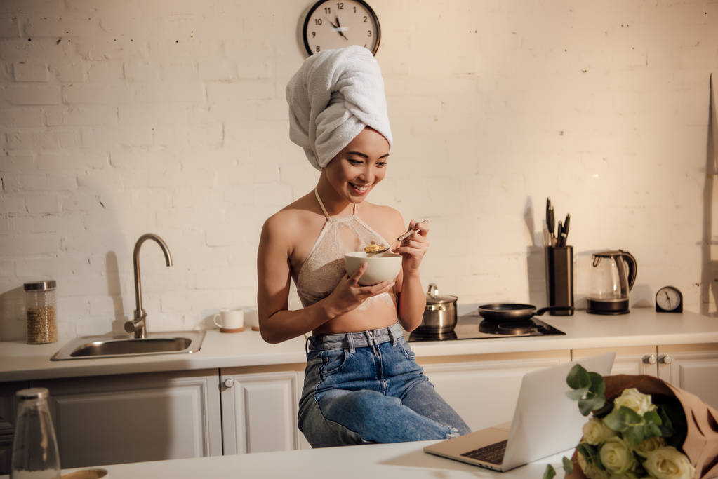 linda menina asiática feliz em laço bralete e toalha na cabeça comer flocos de milho para o café da manhã e usando laptop
 - Foto, Imagem