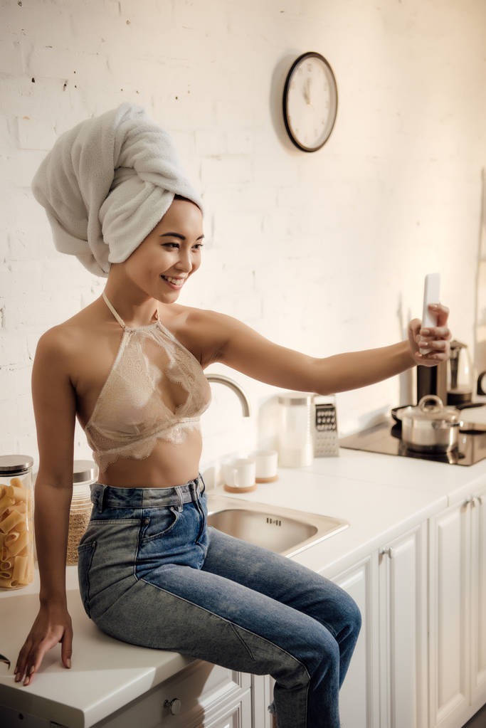 ελκυστικό χαμόγελο νεαρή γυναίκα με πετσέτα στο κεφάλι λαμβάνοντας αυτοπορτρέτα με smartphone στην κουζίνα - Φωτογραφία, εικόνα