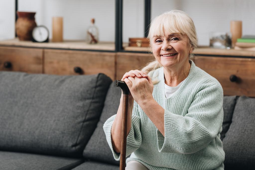 alegre anciana sentada en sofá y sosteniendo bastón
  - Foto, imagen