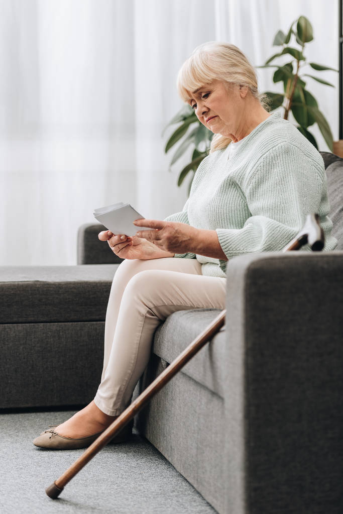 θλιβερή συνταξιούχος γυναίκα με ξανθά μαλλιά, κοιτάζοντας φωτογραφίες ενώ κάθεται στον καναπέ - Φωτογραφία, εικόνα
