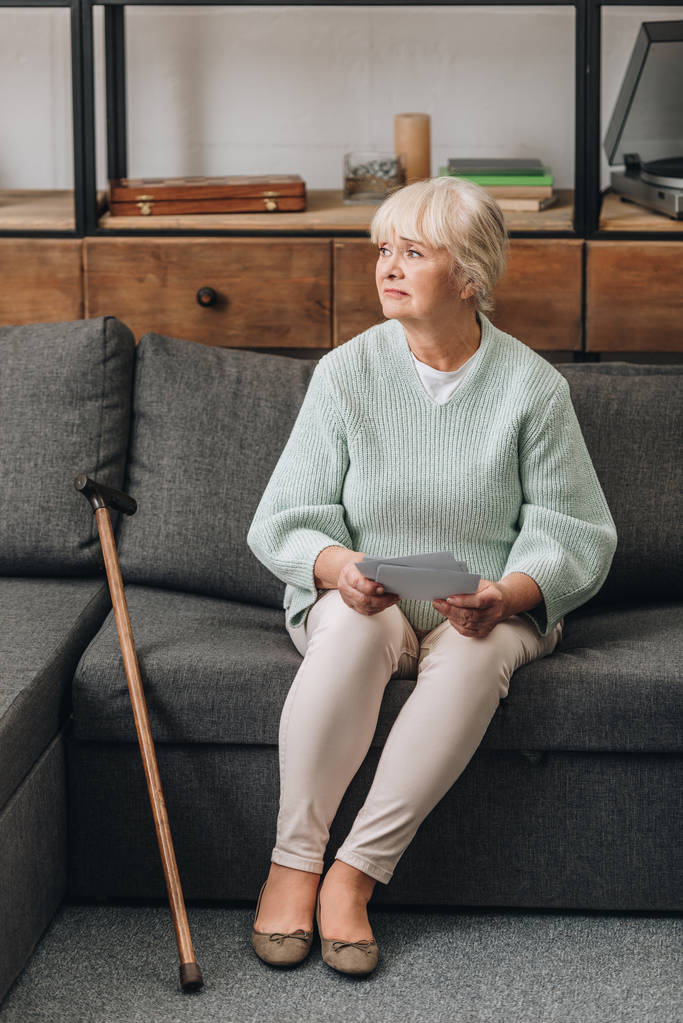Verärgerte Rentnerin mit blonden Haaren hält Fotos hoch, während sie im Wohnzimmer auf dem Sofa sitzt  - Foto, Bild