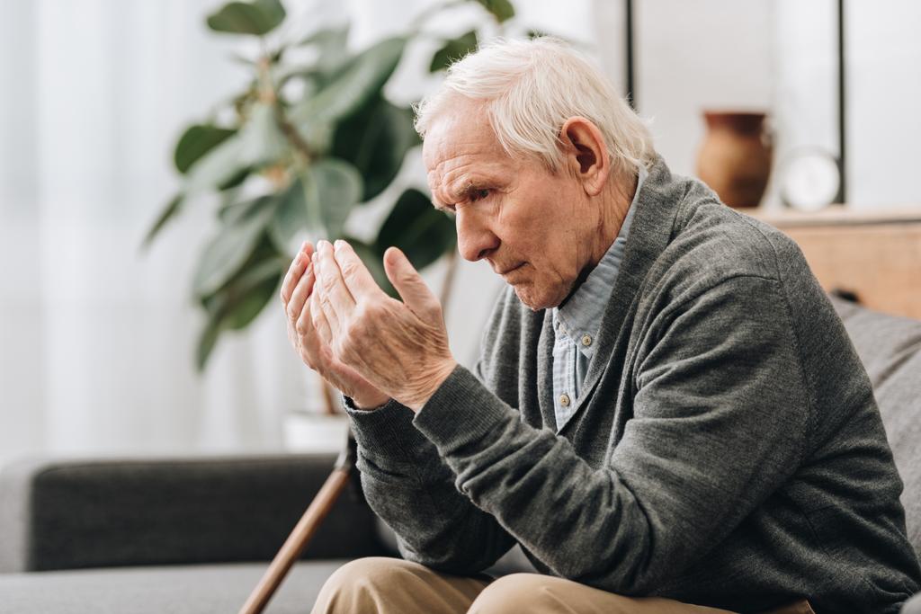  Rentner mit grauen Haaren blickt auf Hände, während er auf dem Sofa sitzt  - Foto, Bild