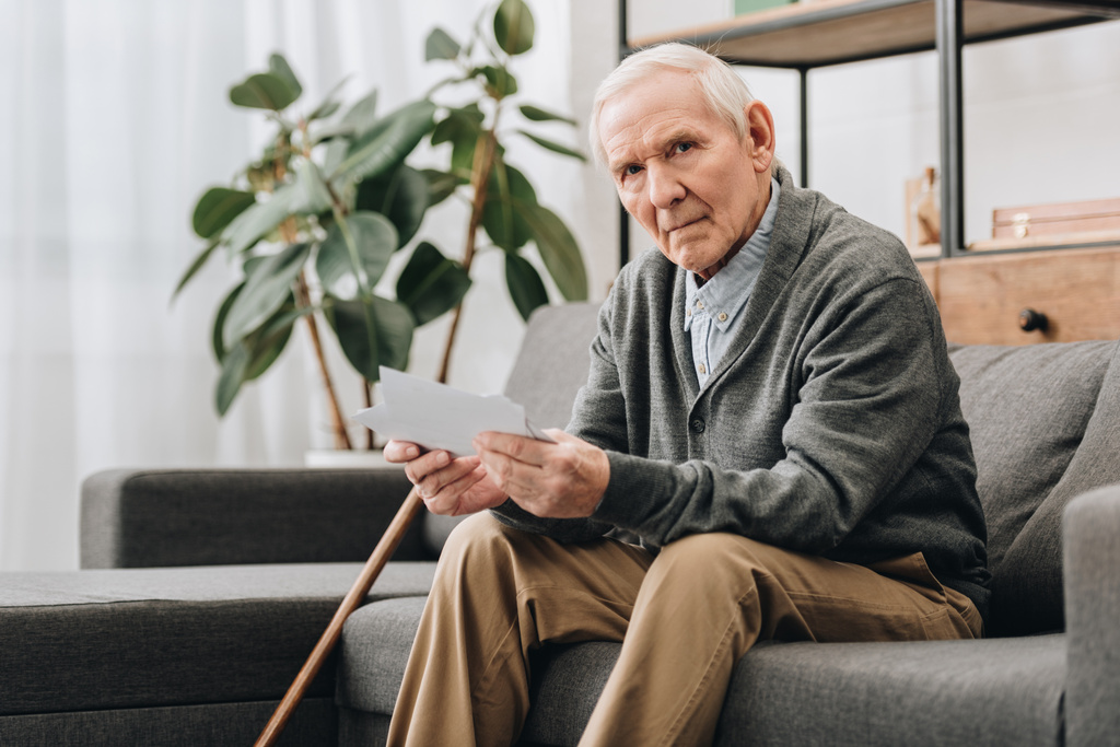 Verärgerter Rentner mit grauen Haaren hält auf Sofa Fotos hoch  - Foto, Bild