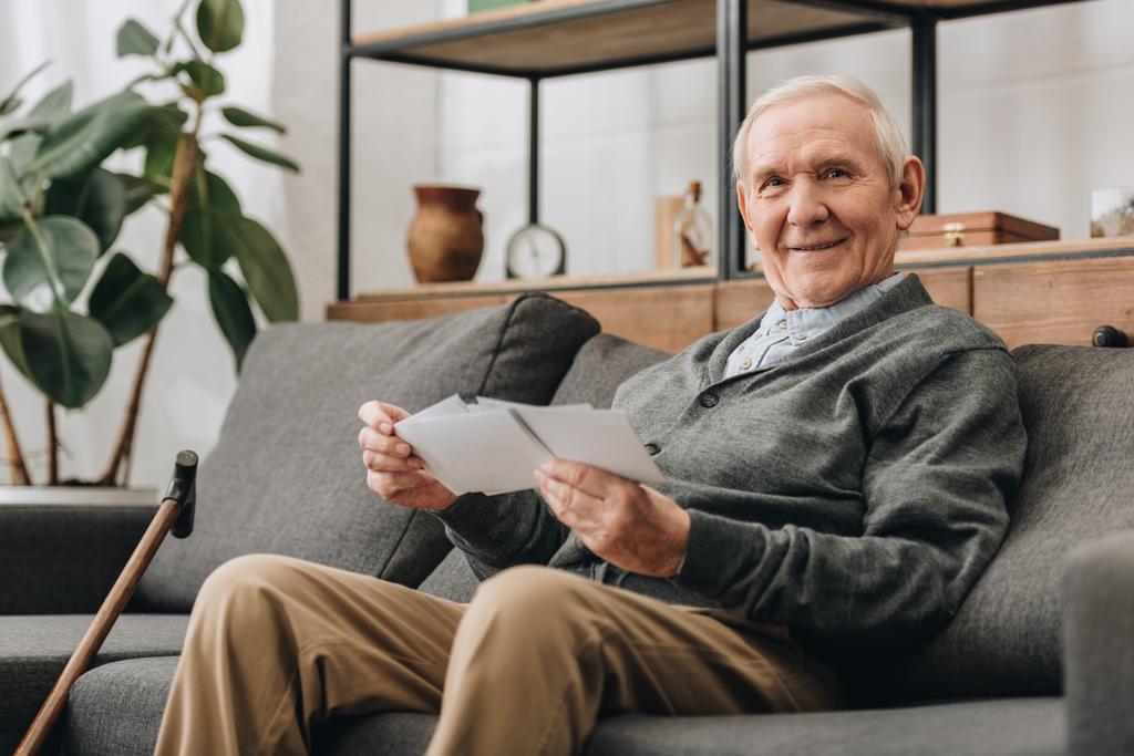 fröhlicher älterer Mann mit grauen Haaren, Fotos in der Hand und auf dem Sofa sitzend - Foto, Bild