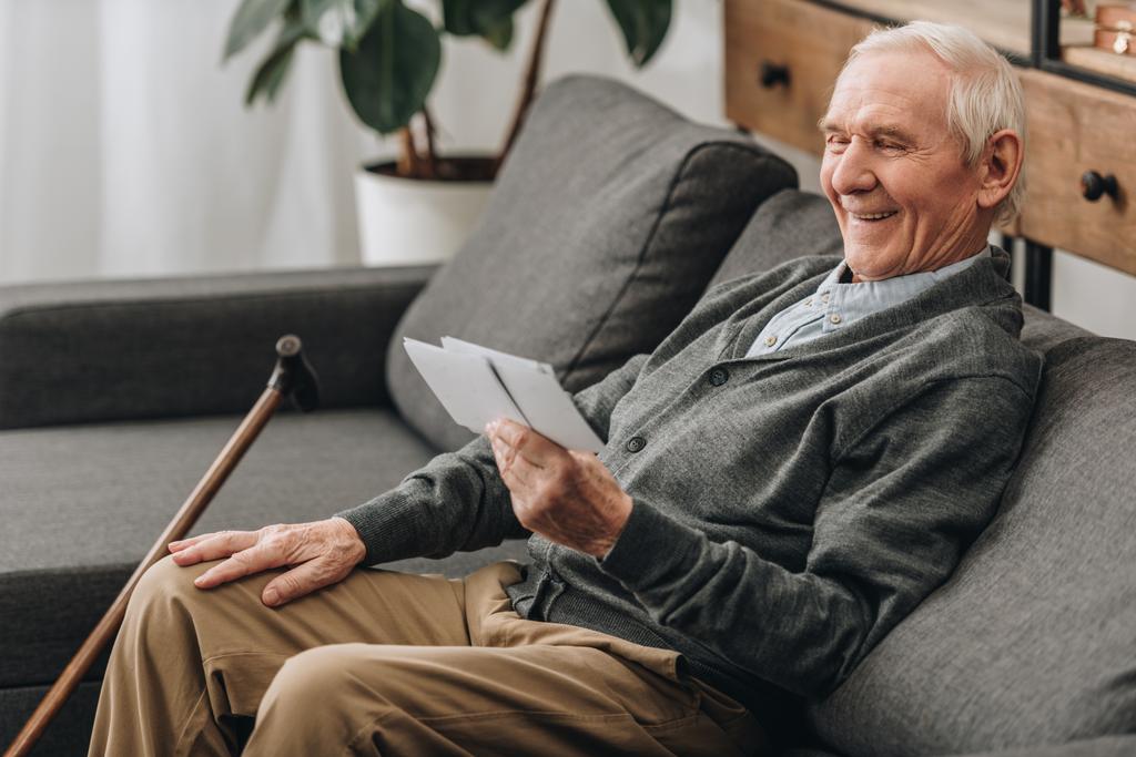 szczęśliwy emeryt z siwe włosy, patrząc na zdjęcia i siedzi na kanapie  - Zdjęcie, obraz