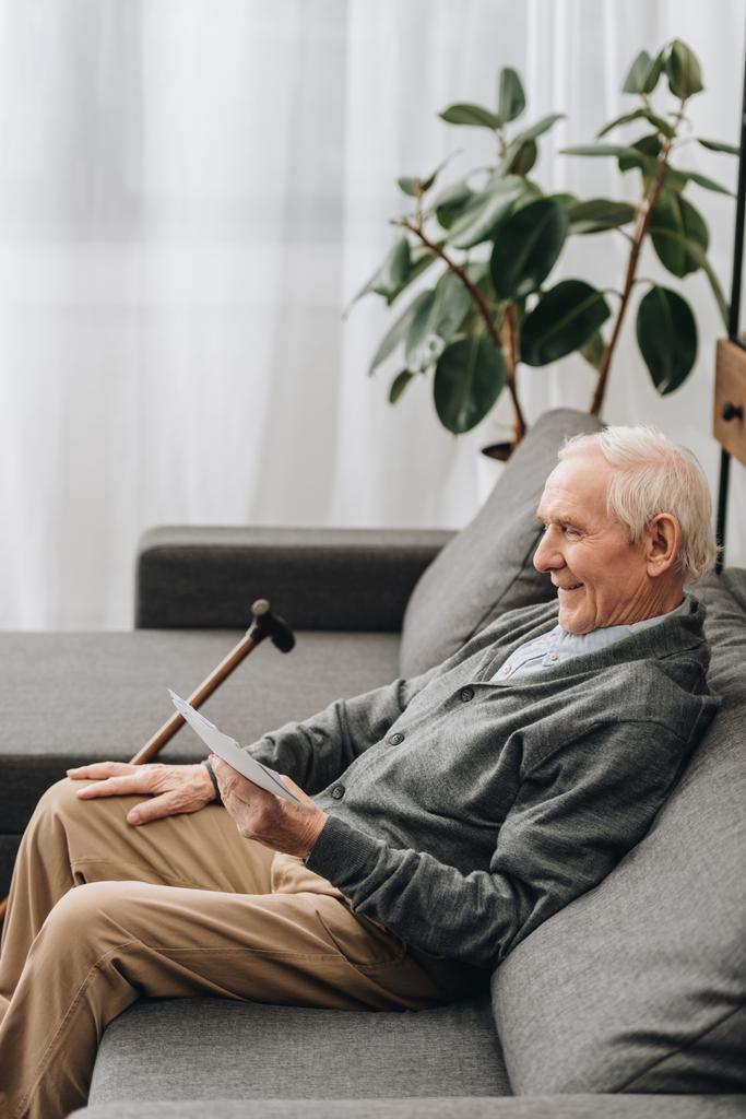 jubilado sonriente con el pelo gris mirando fotos y sentado en el sofá
  - Foto, imagen