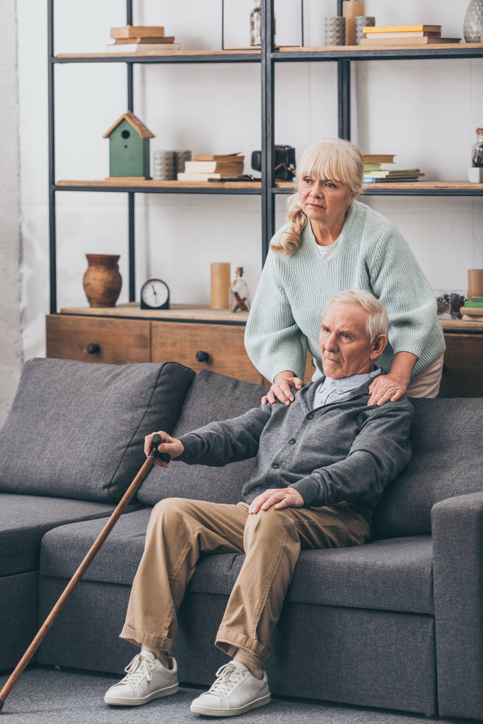Rentnerin umarmt traurigen Ehemann, der auf Sofa im Wohnzimmer sitzt  - Foto, Bild