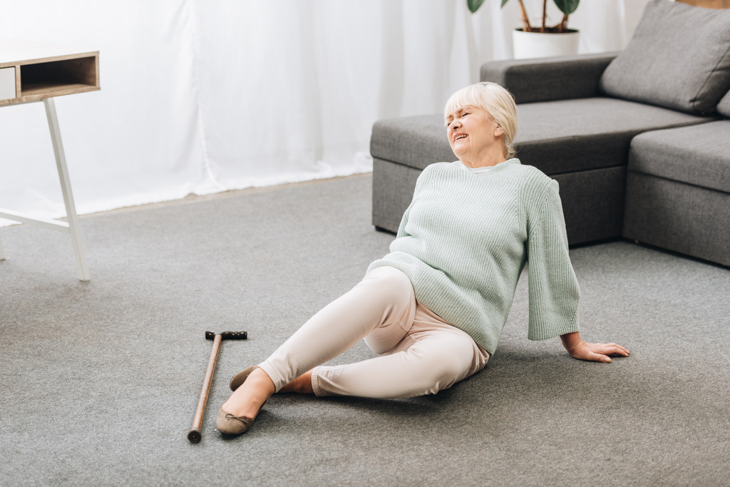 mulher sênior com cabelo loiro deitado no chão perto do sofá e sentindo dor
  - Foto, Imagem