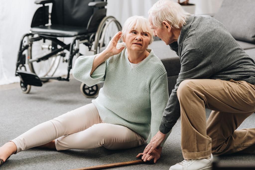 unterstützender pensionierter Ehemann schaut Seniorin auf dem Fußboden sitzend an  - Foto, Bild