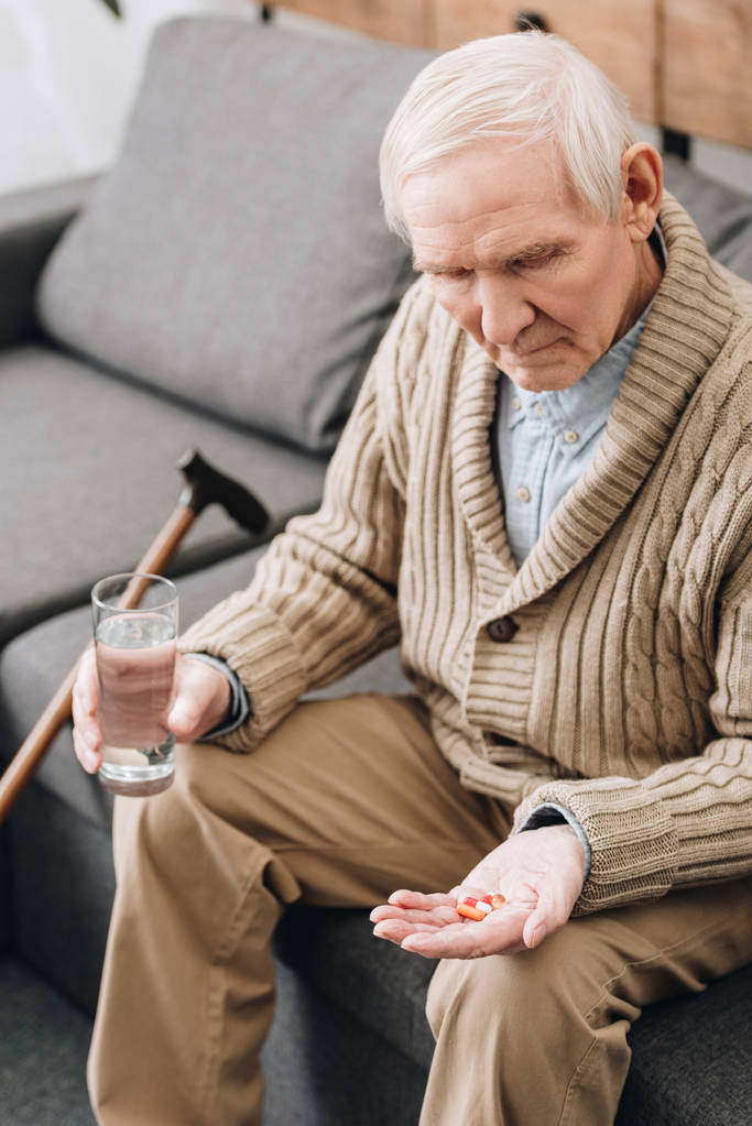 starší muž drží pilulky a sklenici s vodou a při pohledu na dosah ruky - Fotografie, Obrázek