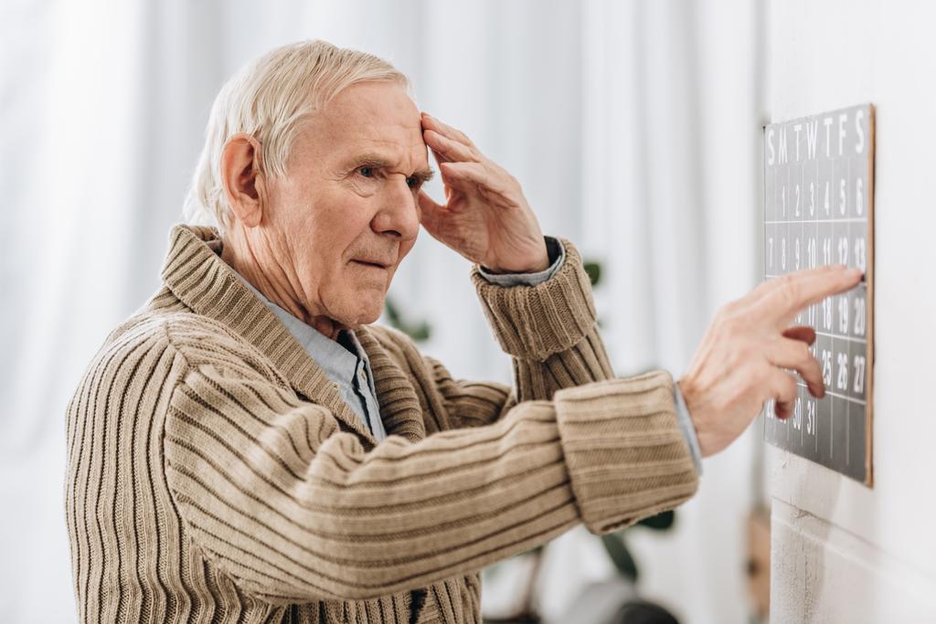 Senior blickt auf Wandkalender und berührt Kopf - Foto, Bild