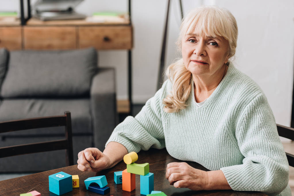 Rentnerin mit blonden Haaren sitzt zu Hause neben Holzspielzeug - Foto, Bild