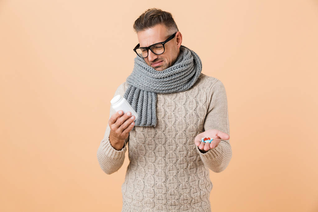Retrato de un hombre molesto vestido con suéter y bufanda de pie aislado sobre fondo beige, mostrando pastillas cápsulas
 - Foto, imagen