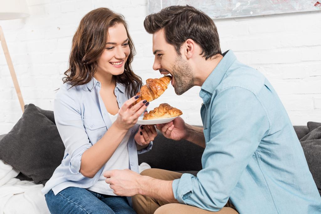 hermosa mujer sonriente alimentando al hombre con croissant durante el desayuno en casa
  - Foto, imagen