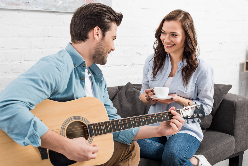 ソファに座って、男の家にアコースティック ギターを演奏しながらコーヒーを飲む女性 - 写真・画像