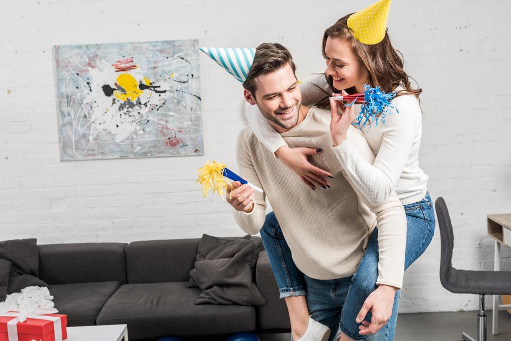 glückliches Paar mit Party-Hörnern und Party-Hüten feiert Geburtstag, während Mann Huckepack-Fahrt zu Frau im Wohnzimmer - Foto, Bild