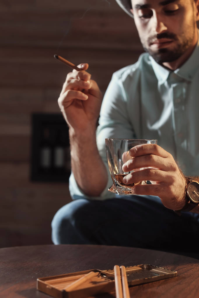Hombre con vaso de whisky y cigarro sentado en la mesa en el interior
 - Foto, imagen