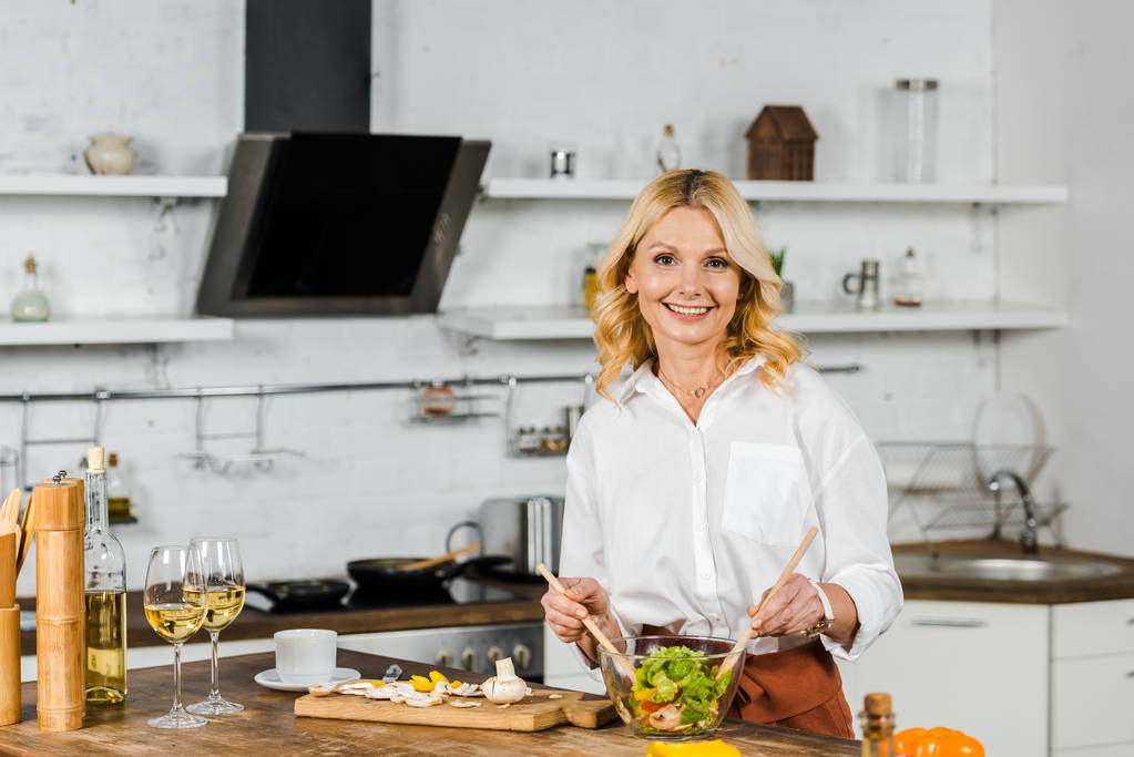 приваблива усміхнена зріла жінка змішує салат на кухні і дивиться на камеру
 - Фото, зображення