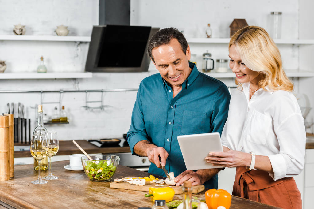 kypsä vaimo käyttäen tabletti ja aviomies leikkaamalla vihanneksia keittiössä
 - Valokuva, kuva