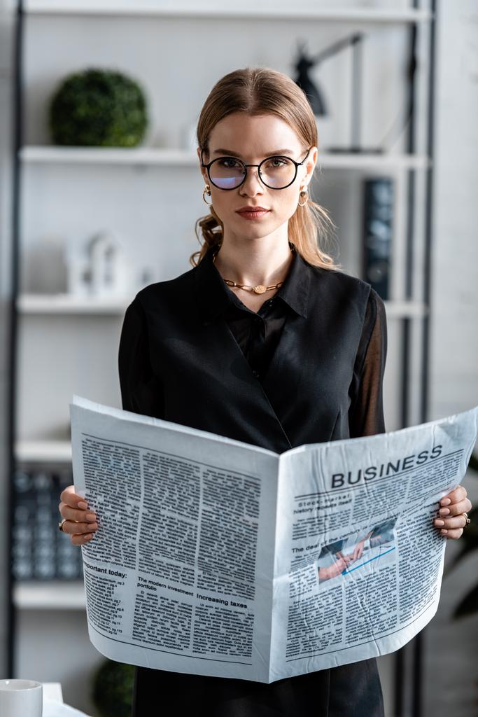 atraktivní podnikatelka v černém oblečení a brýle drží noviny a při pohledu na fotoaparát - Fotografie, Obrázek