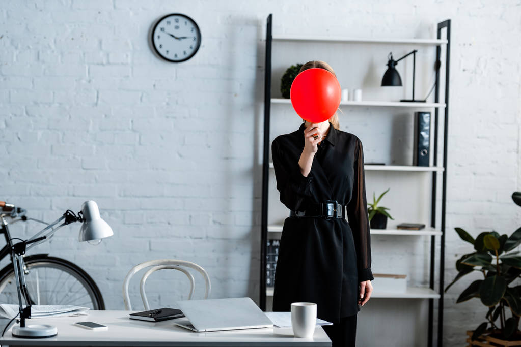 деловая женщина в черной одежде, стоящая возле стола и прячущая лицо за красным воздушным шаром
  - Фото, изображение