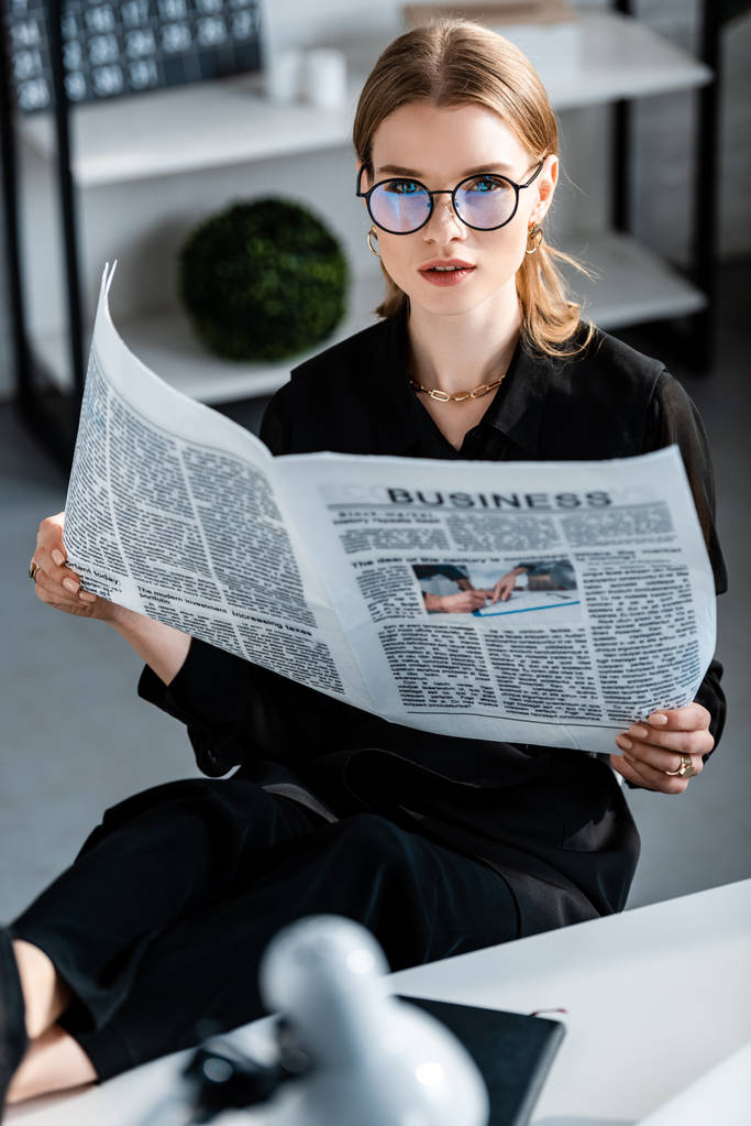 schöne Geschäftsfrau in schwarzer Kleidung und Brille, die am Tisch sitzt und Zeitung hält, während sie in die Kamera schaut - Foto, Bild