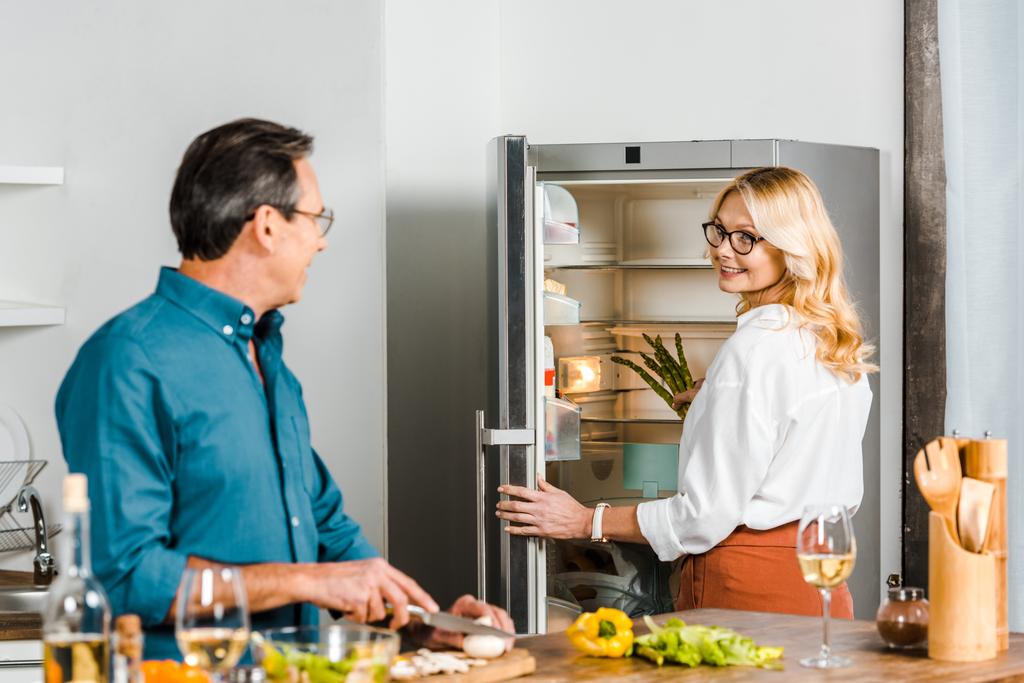 ώριμη γυναίκα παίρνει σπαράγγια από το ψυγείο και ο σύζυγός της κοπής λαχανικών στην κουζίνα - Φωτογραφία, εικόνα