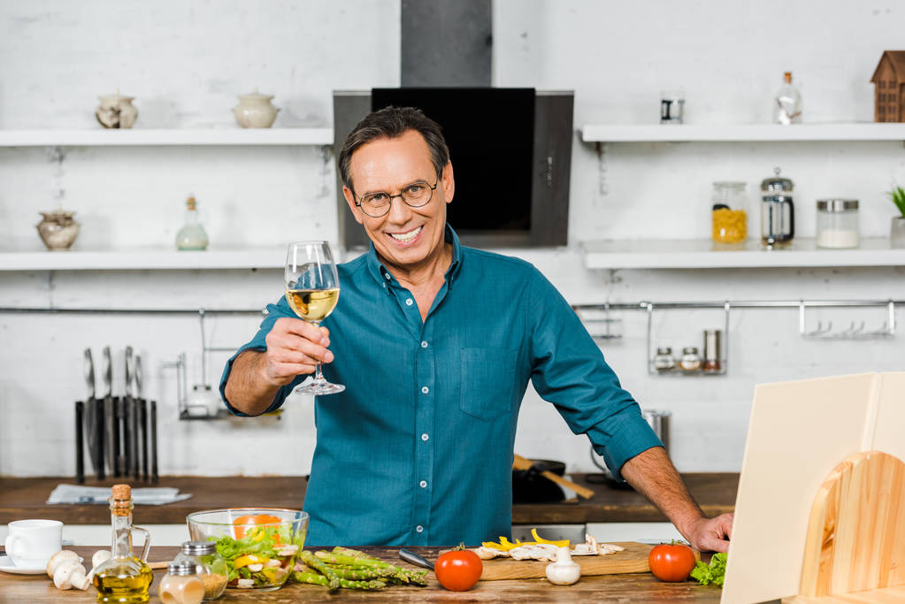 χαμογελώντας όμορφος ώριμος άνδρας δείχνει ποτήρι κρασί στην κουζίνα - Φωτογραφία, εικόνα