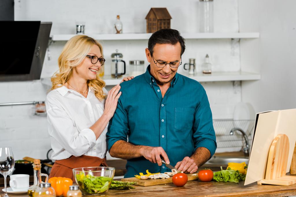 mature femme étreignant mari alors qu'il coupe des légumes dans la cuisine
 - Photo, image