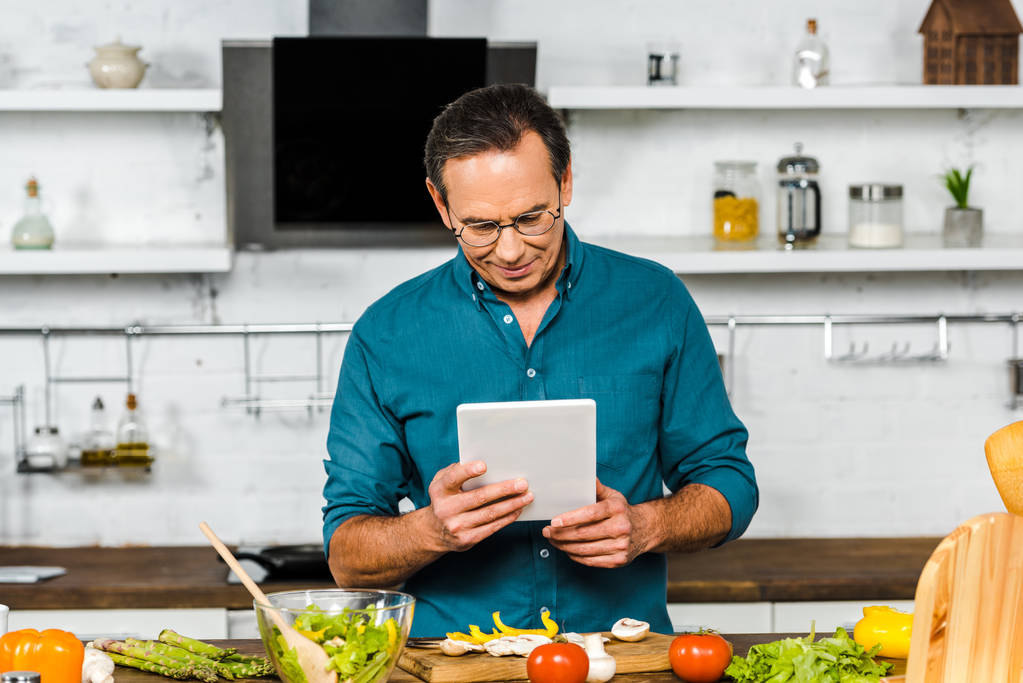guapo hombre maduro usando tableta mientras cocina en la cocina
 - Foto, Imagen