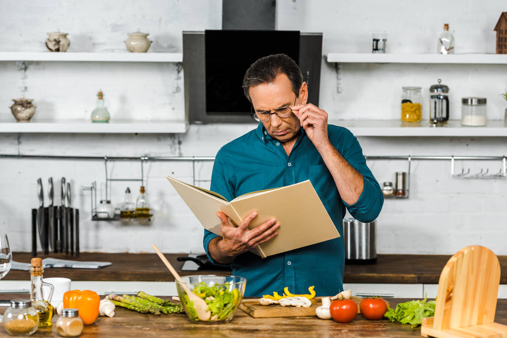 όμορφος ώριμος άνδρας μαγειρέματος στην κουζίνα και βλέπουν το βιβλίο μαγειρικής - Φωτογραφία, εικόνα