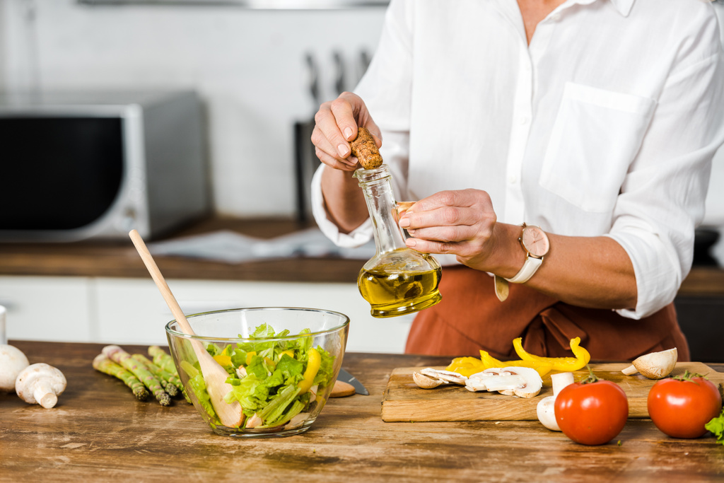 immagine ritagliata di donna matura versando olio in ciotola con insalata in cucina
 - Foto, immagini