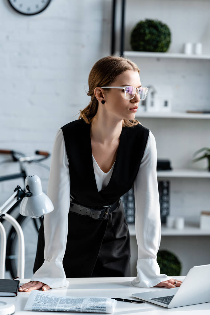 femme d'affaires en tenue formelle et lunettes debout près du bureau avec ordinateur portable et journal sur le lieu de travail
 - Photo, image