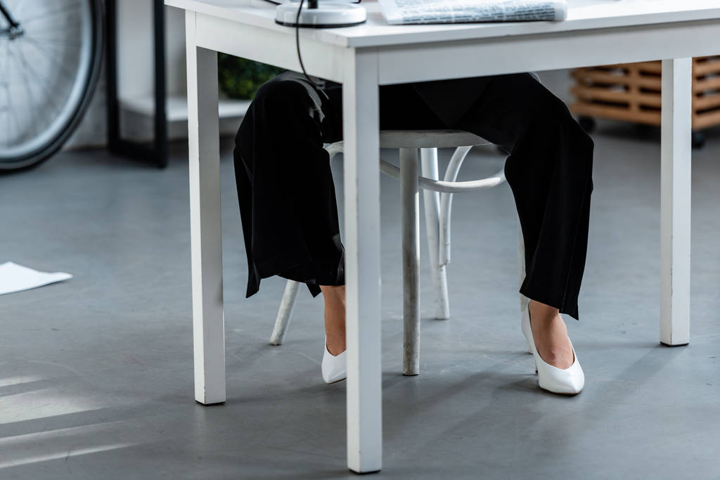 περικοπεί άποψη της γυναίκας σε λευκά παπούτσια και παντελόνια που κάθονται στο τραπέζι στο χώρο εργασίας - Φωτογραφία, εικόνα