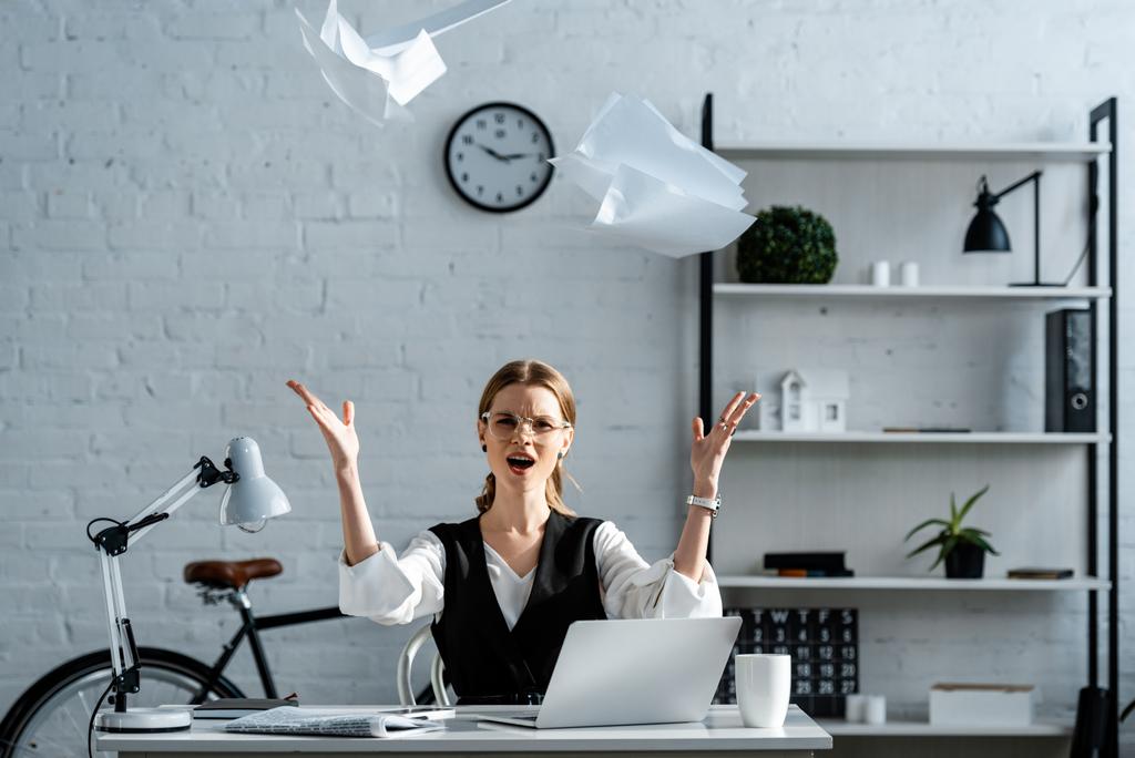 недовольная деловая женщина в формальной одежде сидит за столом и бросает документы в воздух на рабочем месте
 - Фото, изображение