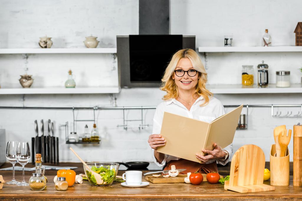улыбающаяся привлекательная зрелая женщина держит книгу рецептов для приготовления пищи на кухне и смотрит в камеру
 - Фото, изображение