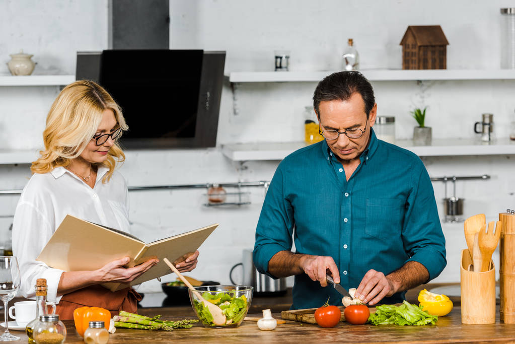 keski-ikäinen vaimo ja aviomies ruoanlaitto yhdessä reseptikirjan kanssa keittiössä
 - Valokuva, kuva